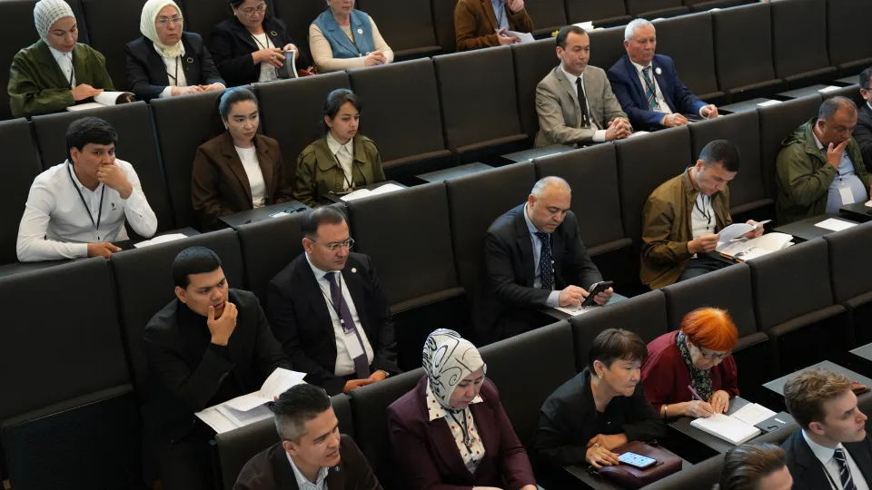 People are seated in a lecture hall, attentively listening and taking notes. The atmosphere is focused and professional, with everyone engaged.