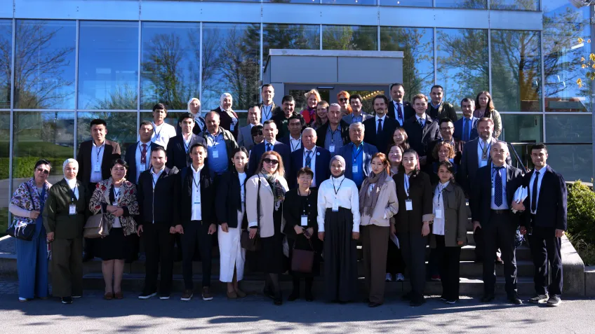 A large group of people stand together on steps outside a modern glass building. They wear professional attire, smiling, suggesting a conference or meeting.
