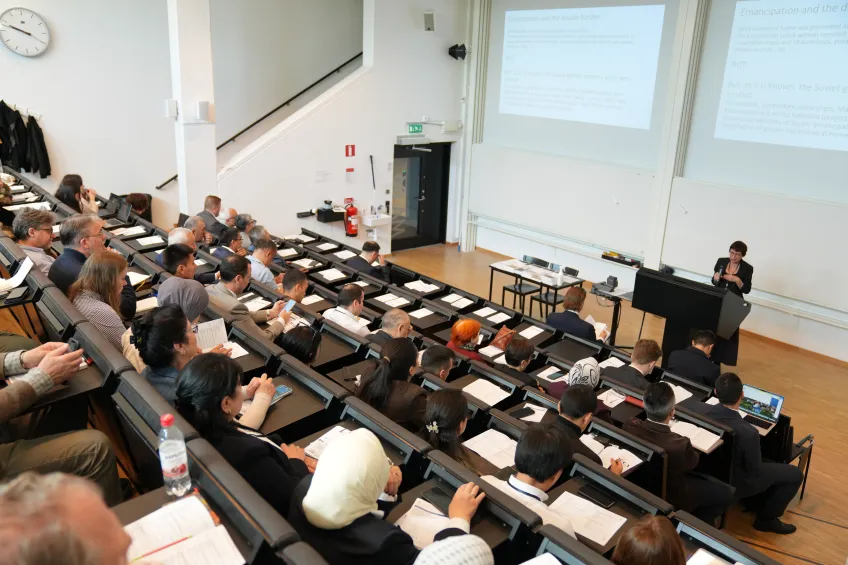 Lecture hall with rows of attendees listening to a speaker at a podium. Projected slides are visible, and the atmosphere is focused and attentive.