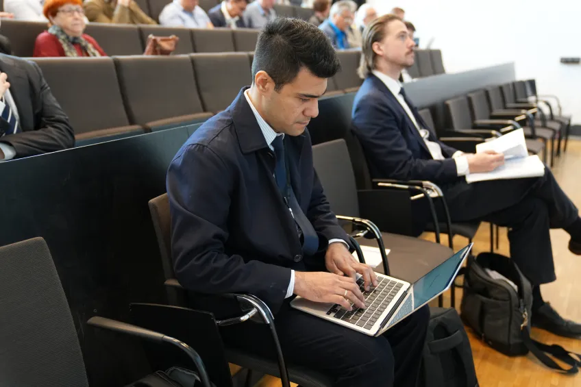 A man in a suit sits in a lecture hall, focused on typing on his laptop. Others in the background take notes, conveying a professional, attentive atmosphere.