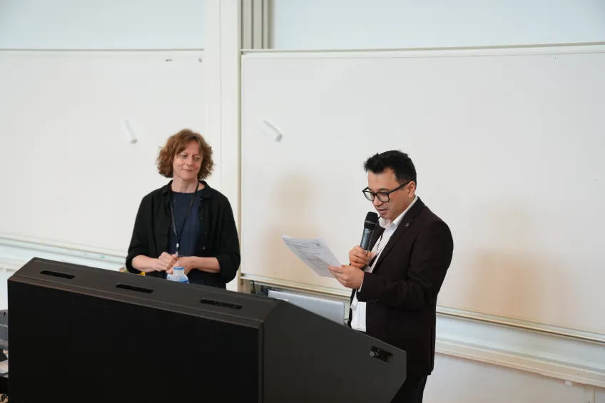 A person speaks into a microphone while holding papers, standing beside a woman in front of a whiteboard. The setting appears to be a lecture or presentation.
