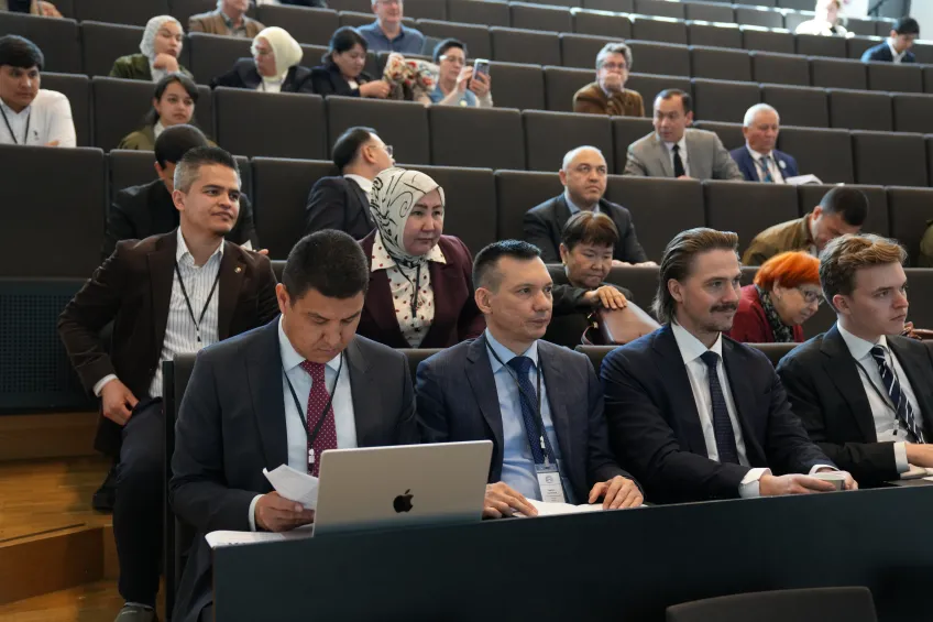 Audience in a conference hall with men and women in suits, some using laptops and taking notes. The setting is formal and attentive.