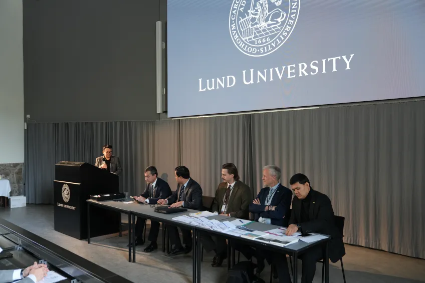 A panel of six individuals sits at a long table with documents at Lund University. A speaker stands at a podium. The setting is formal and academic.