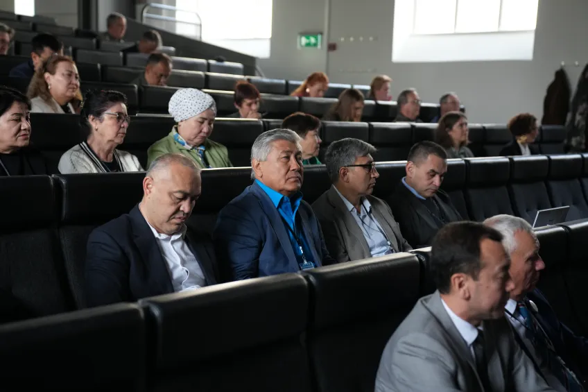 A group of people sits attentively in a dimly lit auditorium. Some take notes, suggesting a serious and focused atmosphere, likely a conference or lecture.