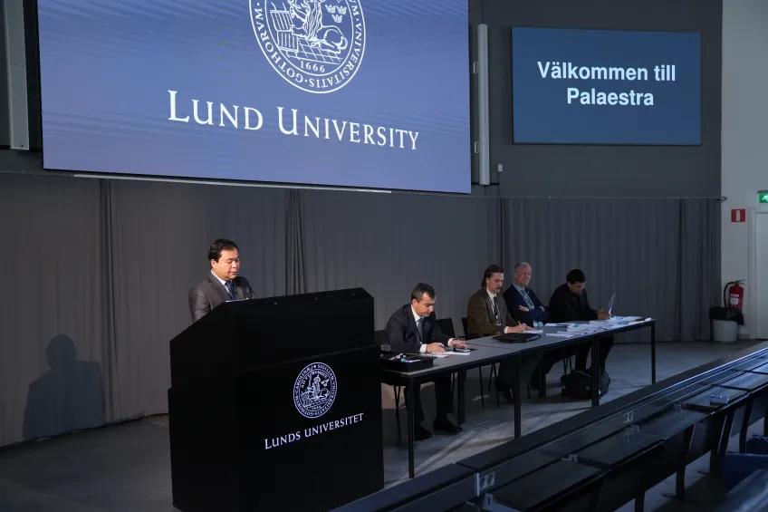 A man speaks at a podium labeled "Lund University," while four men sit at a long table on stage. A screen displays "Välkommen till Palaestra."