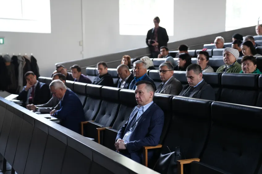 A group of people in formal attire seated in a lecture hall, attentively listening to a presentation. The atmosphere is serious and focused.